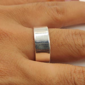 Puede incluir: Primer plano de un anillo plateado con un diseño plano y rectangular. El anillo está hecho de un metal brillante y reflectante y se lleva en un dedo. La banda del anillo mide aproximadamente 0,8 cm de ancho.