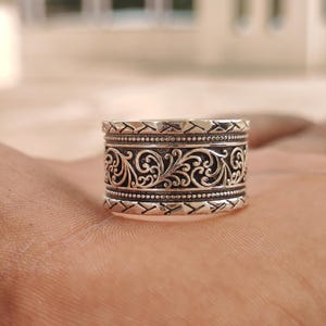 May include: A wide silver band ring featuring a central panel with intricate scrolling vine patterns. The center design is bordered by two rows of beaded detailing, with outer edges displaying a repeating diagonal chevron or slash texture.