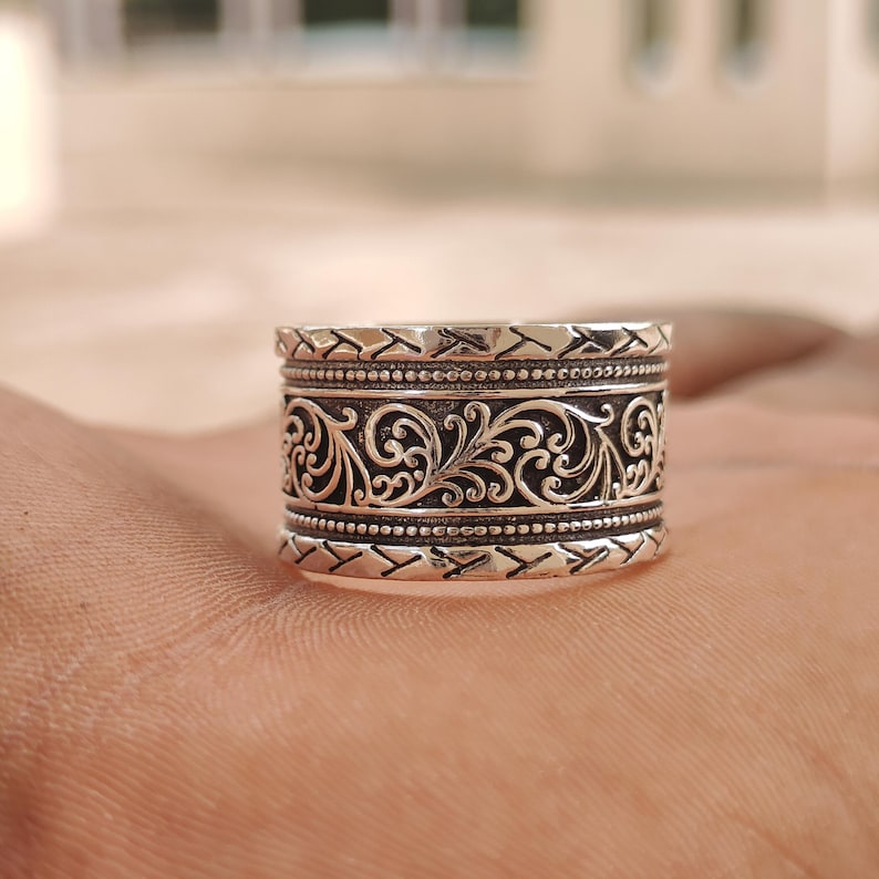 May include: A wide silver band ring featuring a central panel with intricate scrolling vine patterns. The center design is bordered by two rows of beaded detailing, with outer edges displaying a repeating diagonal chevron or slash texture.