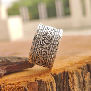 May include: A wide silver ring featuring an intricate, raised floral scrollwork pattern in the center, flanked by two beaded borders and outer bands decorated with a repeating chevron or arrow-like engraved design in a dark oxidized finish.