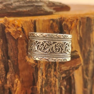 May include: A wide silver band ring featuring an intricate embossed scrollwork pattern in the center, bordered by two rows of beaded detailing and finished with a geometric chevron-style texture along the top and bottom edges of the metal.