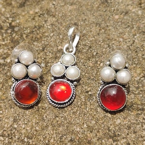 Puede incluir: Un conjunto de joyas con un colgante y pendientes a juego. El colgante y los pendientes tienen un engaste de metal plateado. Cada pieza tiene una gema roja grande y redonda en la parte inferior, con tres perlas blancas encima. Los pendientes tienen respaldos transparentes.