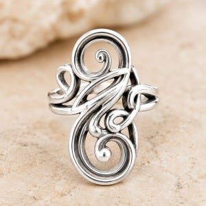 May include: A polished silver ring featuring an intricate openwork design composed of multiple swirling, curved metal lines that form a continuous, elongated pattern. The metal surface has darkened recessed areas to highlight the scrollwork details.
