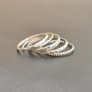May include: A set of five silver-toned rings stacked together. Each ring features a distinct texture, including smooth, hammered, zigzag, cross-hatched, and twisted rope patterns. The rings are arranged in a cascading row, showcasing their metallic finish.