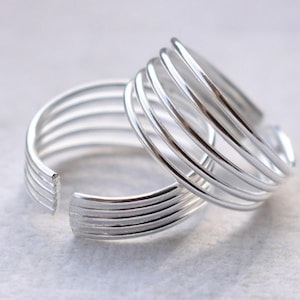 May include: Two silver toe rings. Each ring features a series of parallel, curved bands. The rings are open at the front, allowing for adjustment. The rings are displayed on a light gray surface.