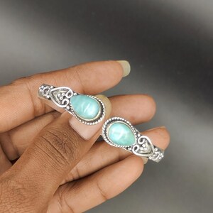 Puede incluir: Dos anillos de plata con piedras preciosas en forma de lágrima de color azul claro. Los anillos tienen detalles ornamentales de estilo filigrana alrededor de las piedras preciosas y en las bandas. Las piedras están engastadas en un engaste de bisel. Fondo oscuro.