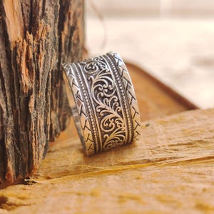 May include: A wide silver ring featuring an intricate embossed floral scroll pattern in the center, bordered by two thin beaded lines and outer bands with a repeating chevron-like texture. The metal has a dark oxidized finish in the recessed areas.