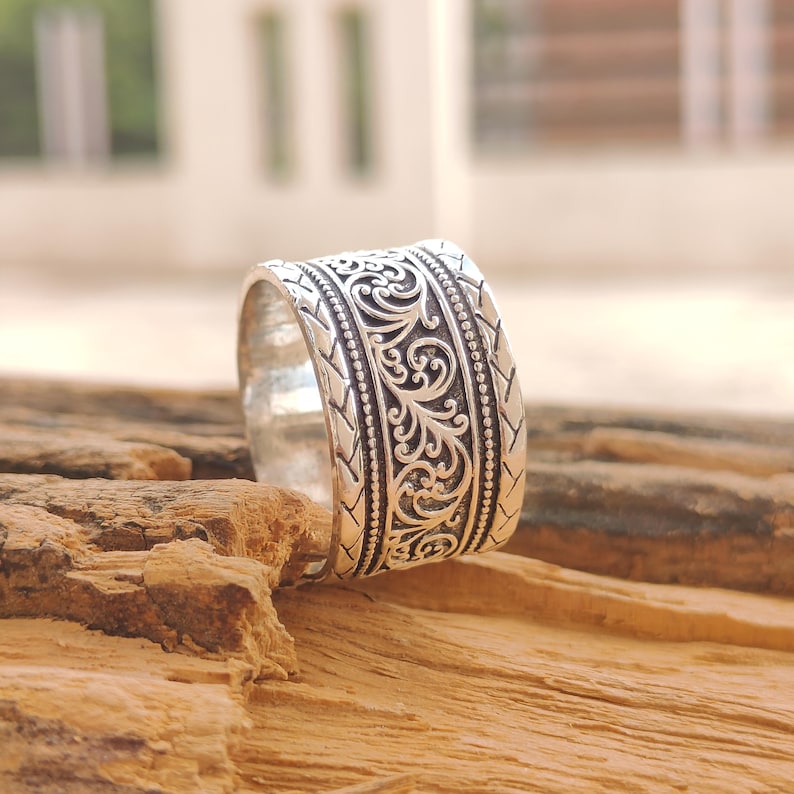 May include: A wide silver band ring featuring an intricate central scrollwork pattern flanked by beaded borders and outer edges with a repeating chevron-style engraved design. The metal has a polished finish with darkened recessed areas.