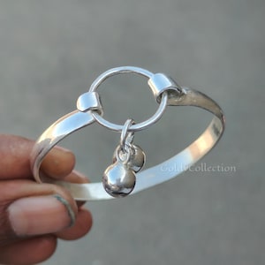 Op de afbeelding: Een zilverkleurige armband met een cirkelvormig ontwerp en twee hangende bolvormige bedels. De armband heeft een gepolijste afwerking. De tekst "Gold Collection" is zichtbaar op de armband.