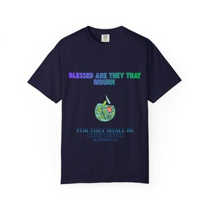 Op de afbeelding: Een marineblauw t-shirt met korte mouwen met de tekst BLESSED ARE THEY THAT MOURN in een blauw-groen verloop, een afbeelding van een kleine kom met een bloem en bladeren, en de tekst FOR THEY SHALL BE COMFORTED MATTHEW 5:5 in het blauw.