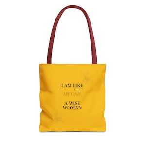 May include: A yellow tote bag with dark red handles. The front features black text that reads I AM LIKE ABIGAIL A WISE WOMAN, with a faint gold-colored floral graphic design visible in the background behind the printed text.