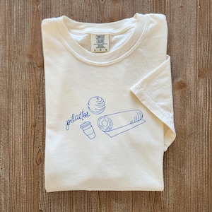 May include: Cream-colored t-shirt with a blue Pilates-themed graphic. The design includes the word "pilates" in cursive, a ball, a rolled mat, and a coffee cup. The shirt is folded and has a tag that says "Comfort Colors".