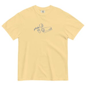 May include: A light yellow t-shirt with a crew neck. The shirt features a blue line drawing of a Pilates ball, a coffee cup, and a rolled mat. The word "pilates" is written in cursive above the illustrations.
