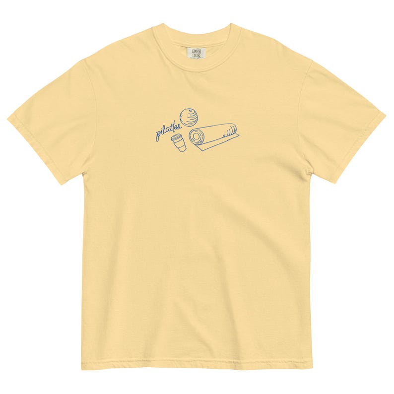 May include: A light yellow t-shirt with a crew neck. The shirt features a blue line drawing of a Pilates ball, a coffee cup, and a rolled mat. The word "pilates" is written in cursive above the illustrations.