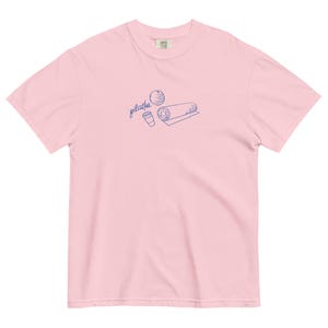 May include: A light pink t-shirt with a blue line drawing of a Pilates ball, a rolled mat, and a coffee cup. The word "pilates" is written in cursive above the objects. The shirt is made of a soft material.