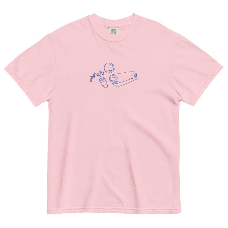 May include: A light pink t-shirt with a blue line drawing of a Pilates ball, a rolled mat, and a coffee cup. The word "pilates" is written in cursive above the objects. The shirt is made of a soft material.