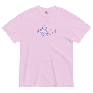 May include: A light pink t-shirt with a blue graphic of a pilates ball, a rolled mat, and a coffee cup. The word "pilates" is written in cursive above the graphic. The shirt is made of a soft material.
