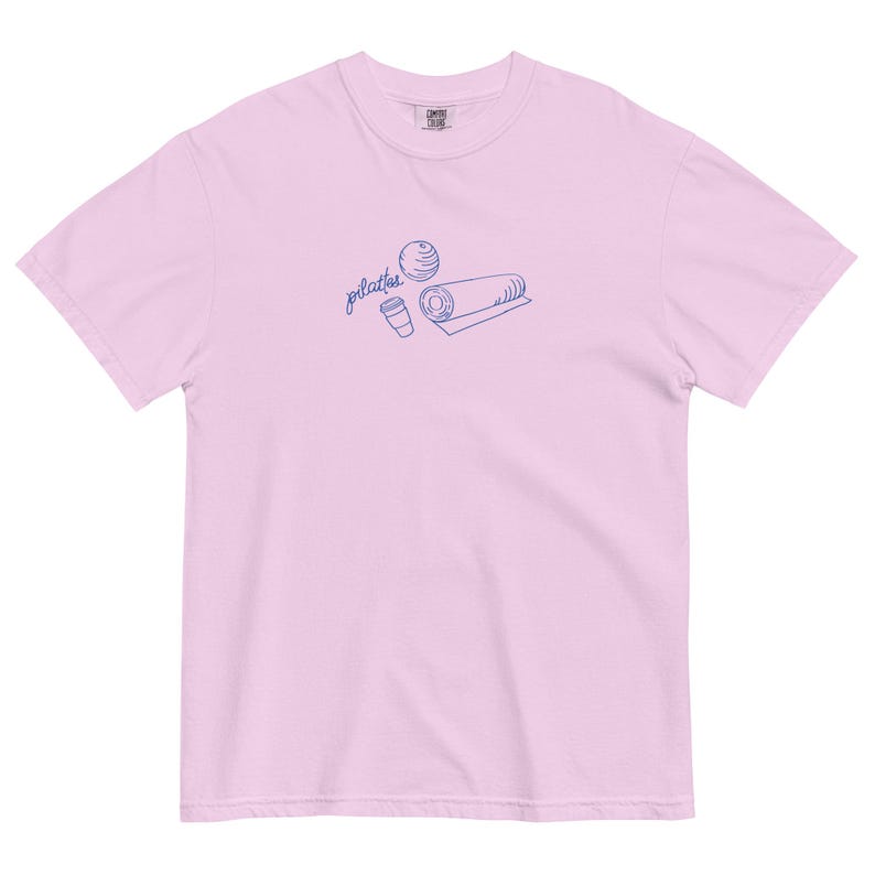 May include: A light pink t-shirt with a blue graphic of a pilates ball, a rolled mat, and a coffee cup. The word "pilates" is written in cursive above the graphic. The shirt is made of a soft material.