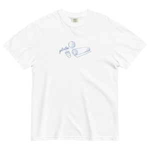 May include: White t-shirt with a blue line drawing of pilates equipment, including a ball, a mat, and a coffee cup. The word "pilates" is written in cursive above the illustrations. The shirt is made of a soft, comfortable material.