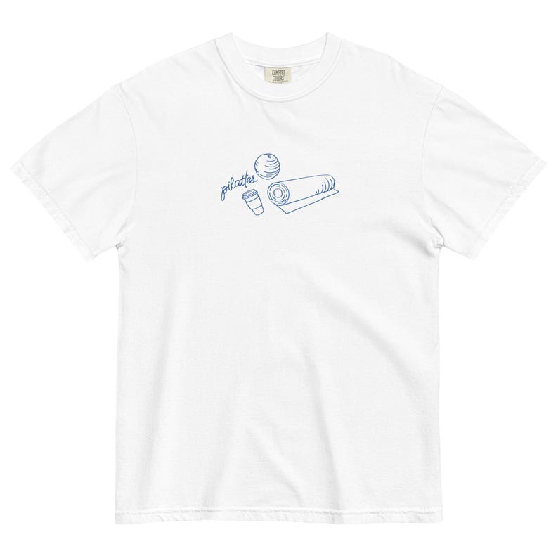 May include: White t-shirt with a blue line drawing of pilates equipment, including a ball, a mat, and a coffee cup. The word "pilates" is written in cursive above the illustrations. The shirt is made of a soft, comfortable material.