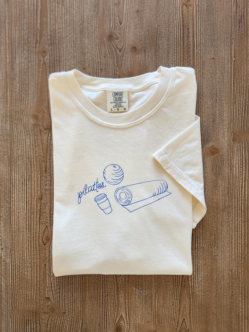 May include: Cream-colored t-shirt with a blue Pilates-themed graphic. The design includes the word "pilates" in cursive, a ball, a rolled mat, and a coffee cup. The shirt is folded and has a tag that says "Comfort Colors".
