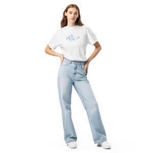 May include: A white t-shirt with a blue line drawing of a tube of paint, a paint palette, and a paintbrush. The model is wearing light blue jeans and white sneakers.