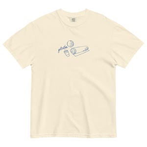 May include: Cream-colored t-shirt with a blue line drawing of pilates equipment: a ball, a rolled mat, and a coffee cup. The word "pilates" is written in cursive above the illustrations. The shirt is made of a soft, comfortable material.