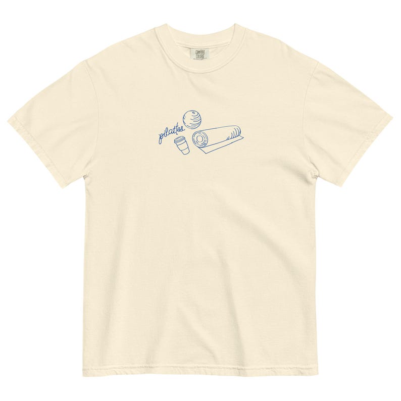 May include: Cream-colored t-shirt with a blue line drawing of pilates equipment: a ball, a rolled mat, and a coffee cup. The word "pilates" is written in cursive above the illustrations. The shirt is made of a soft, comfortable material.