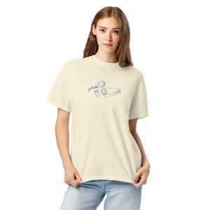 May include: Cream-colored t-shirt with a blue line drawing of a yoga mat, coffee cup, and ball. The word "gelatful" is written above the drawing in a script font. The model is wearing blue jeans.