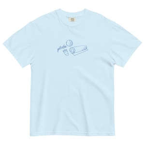 May include: Light blue t-shirt with a blue line drawing of a Pilates ball, a rolled mat, and a coffee cup. The word "pilates" is written in cursive above the illustrations. The shirt is made of a soft, comfortable material.