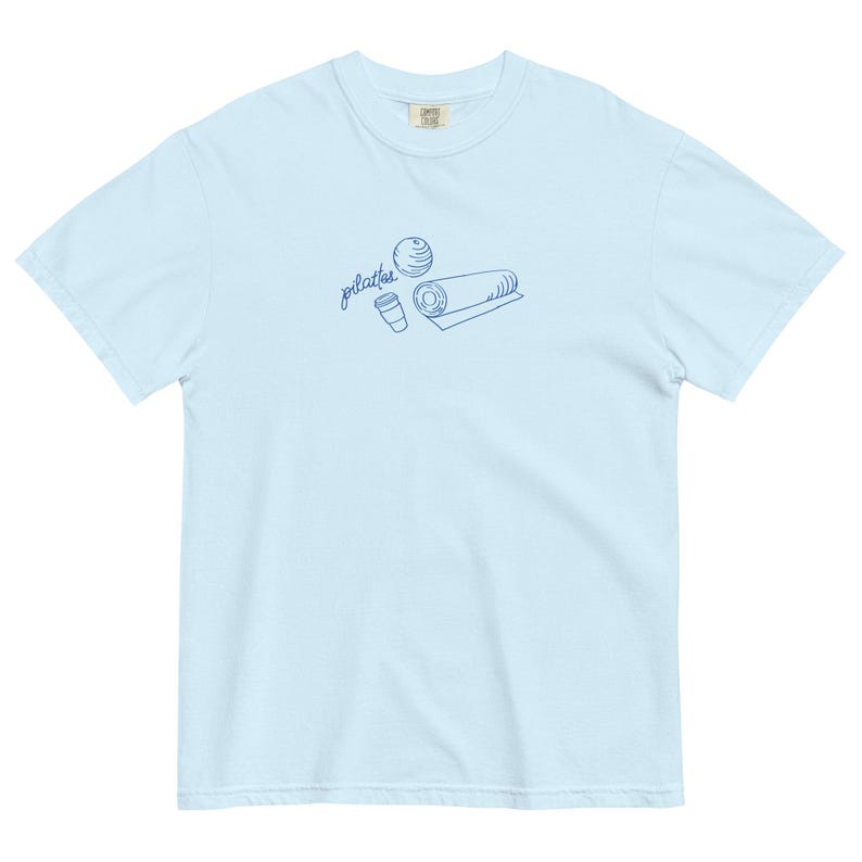 May include: Light blue t-shirt with a blue line drawing of a Pilates ball, a rolled mat, and a coffee cup. The word "pilates" is written in cursive above the illustrations. The shirt is made of a soft, comfortable material.
