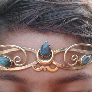 May include: A gold-coloured metal crown with a central, teardrop-shaped blue gemstone and smaller matching stones. The crown has an ornate, swirling design, suitable for special occasions or costume use.