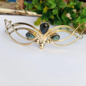 Op de afbeelding: Een goudkleurige metalen haarband met een gebogen ontwerp. Het heeft drie blauwgroene edelsteenaccenten. De haarband heeft een open, vloeiend ontwerp, met een centrale, grotere edelsteen en twee kleinere.