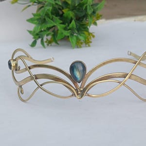 Peut inclure: Un diadème en métal doré avec un design complexe et fluide. La couronne comporte trois pierres sombres et une pierre centrale irisée en forme de goutte. Le design est élégant et adapté aux occasions spéciales.