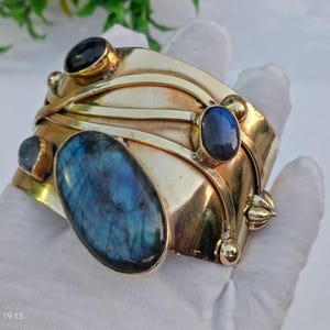 May include: A wide, gold-toned metal cuff bracelet with an oval, blue labradorite stone and several smaller, dark blue stones. The bracelet has an intricate design with curved metal bands and decorative accents.