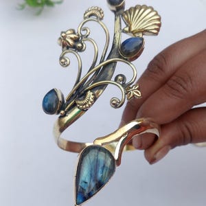 Könnte beinhalten: Ein Armreif aus Messing mit floralem und Muscheldesign, besetzt mit blauen Labradorit-Edelsteinen. Der Armreif hat einen einzigartigen Stil mit einem großen tropfenförmigen Stein an der Basis. Der Armreif wird von einer Hand gehalten.