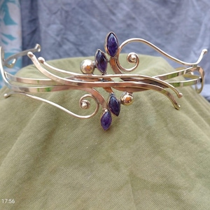 May include: A silver-toned metal tiara featuring flowing, curved wire designs and small, polished spherical accents. The headpiece is adorned with five marquise-cut, deep purple gemstones arranged in clusters along the center of the band.