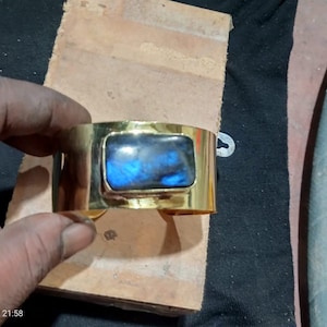 May include: A wide, polished gold-tone metal cuff bracelet featuring a large, rectangular labradorite gemstone set in the center. The stone displays a prominent iridescent blue flash against a dark, mottled gray background within the setting.