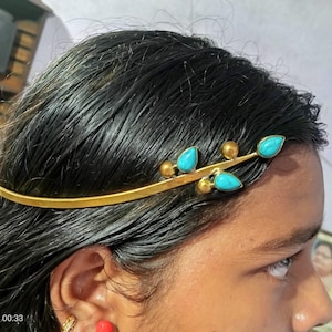 Peut inclure: Un serre-tête en métal doré au design en forme de branche, orné de trois pierres turquoise en forme de goutte et de deux petites sphères dorées. L'accessoire repose sur le front, présentant une bande incurvée lisse et des éléments décoratifs.