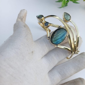 May include: A gold-toned metal cuff bracelet featuring three oval-shaped labradorite stones and two smaller round stones. The bracelet has a unique, open design with curved lines and is set against a white background.