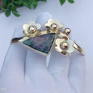 Puede incluir: Un brazalete de metal dorado con una piedra triangular de patrones moteados en rosa y verde oscuro. El brazalete está adornado con tres formas de flores doradas pulidas, cada una con un acento esférico central, dispuestas a lo largo.