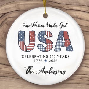 May include: A white ceramic ornament with a gold string for hanging. The ornament features the text "One Nation Under God" and "USA" in a patchwork design of the American flag. It celebrates 250 years.