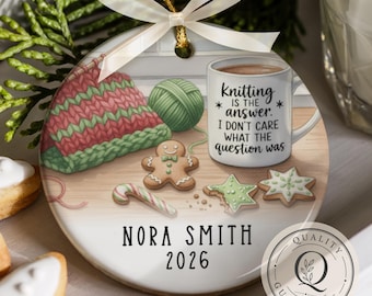 May include: A round ceramic ornament featuring a graphic of a knitted piece, a green yarn ball, a gingerbread man, a candy cane, two star cookies, and a mug with the text Knitting is the answer. I don't care what the question was. Nora Smith 2026.
