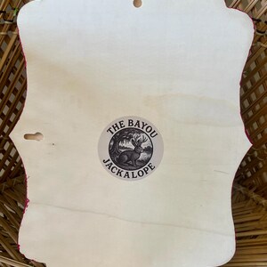 May include: A wooden wall hanging with a decorative edge, featuring a light-colored wood surface. The back of the piece has a small circular sticker with the text "THE BAYOU JACKALOPE" and an illustration of a rabbit. The edges are trimmed with a pink fabric.