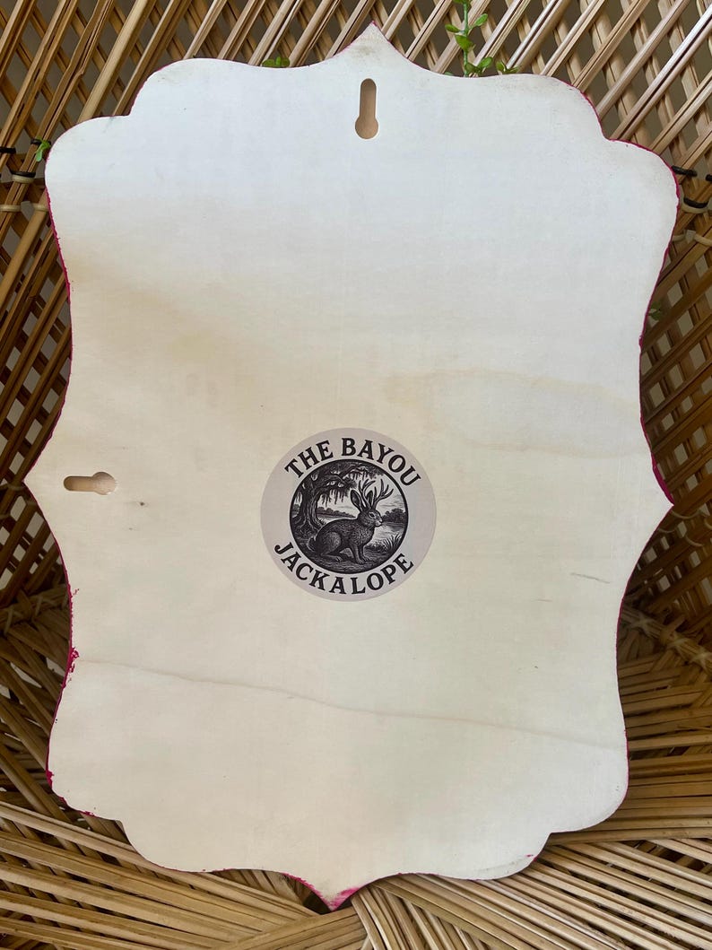 May include: A wooden wall hanging with a decorative edge, featuring a light-colored wood surface. The back of the piece has a small circular sticker with the text "THE BAYOU JACKALOPE" and an illustration of a rabbit. The edges are trimmed with a pink fabric.
