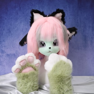 May include: A furry character head and paws. The head features pink hair, black and white ears, and a green face. The paws are green and white with pink pads. The character is set against a blue backdrop.