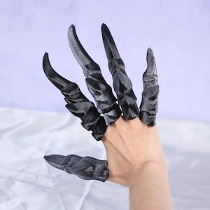 May include: Black claw-shaped finger extensions. The extensions are worn on the fingers and appear to be made of a hard, textured material. The claws are curved and pointed, giving a menacing appearance. The hand is against a light purple background.