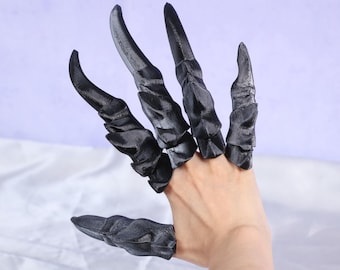 May include: Black claw-shaped finger extensions. The extensions are worn on the fingers and appear to be made of a hard, textured material. The claws are curved and pointed, giving a menacing appearance. The hand is against a light purple background.