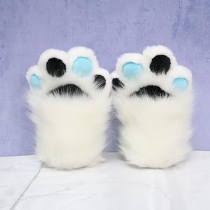 May include: A pair of white faux fur paw gloves. Each paw has black and light blue pads. The gloves are designed to resemble animal paws, with a fluffy, soft appearance. The background is a light purple color.