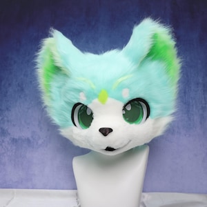 Peut inclure: Tête de renard en fourrure, stylisée, avec de grands yeux verts expressifs. La tête est principalement bleu clair avec des accents verts sur les oreilles et un museau blanc. Un petit losange vert orne le front. La tête est montée sur un mannequin blanc.